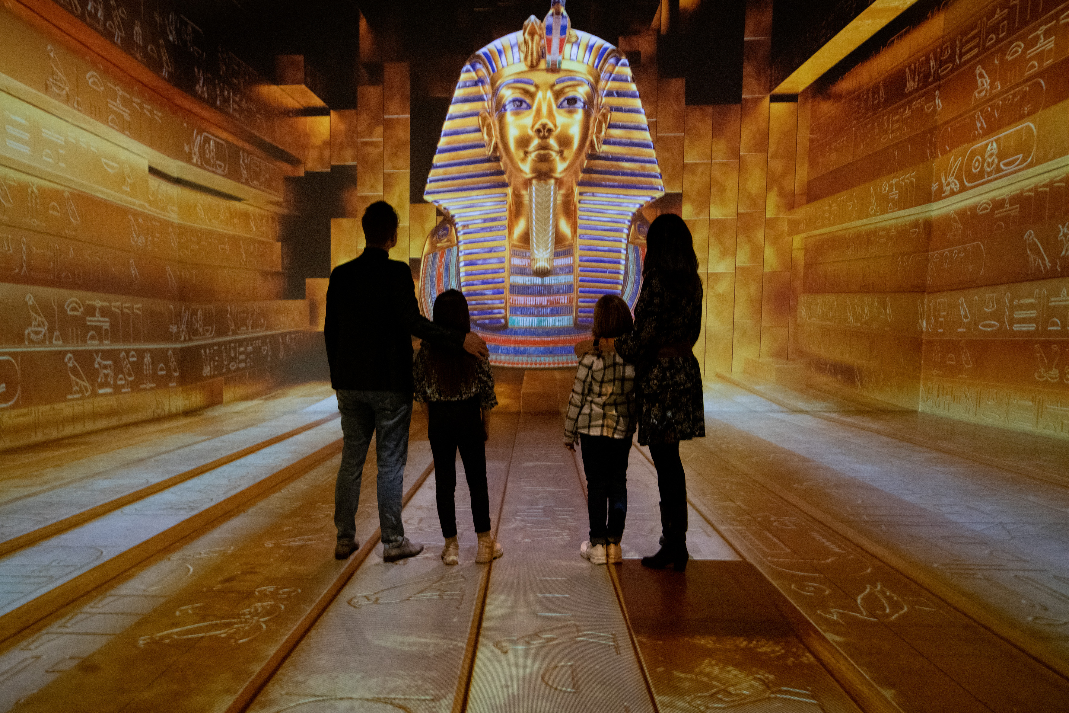 A group of four people stands facing an illuminated depiction of Tutankhamun's golden mask in a corridor adorned with ancient Egyptian hieroglyphics.
